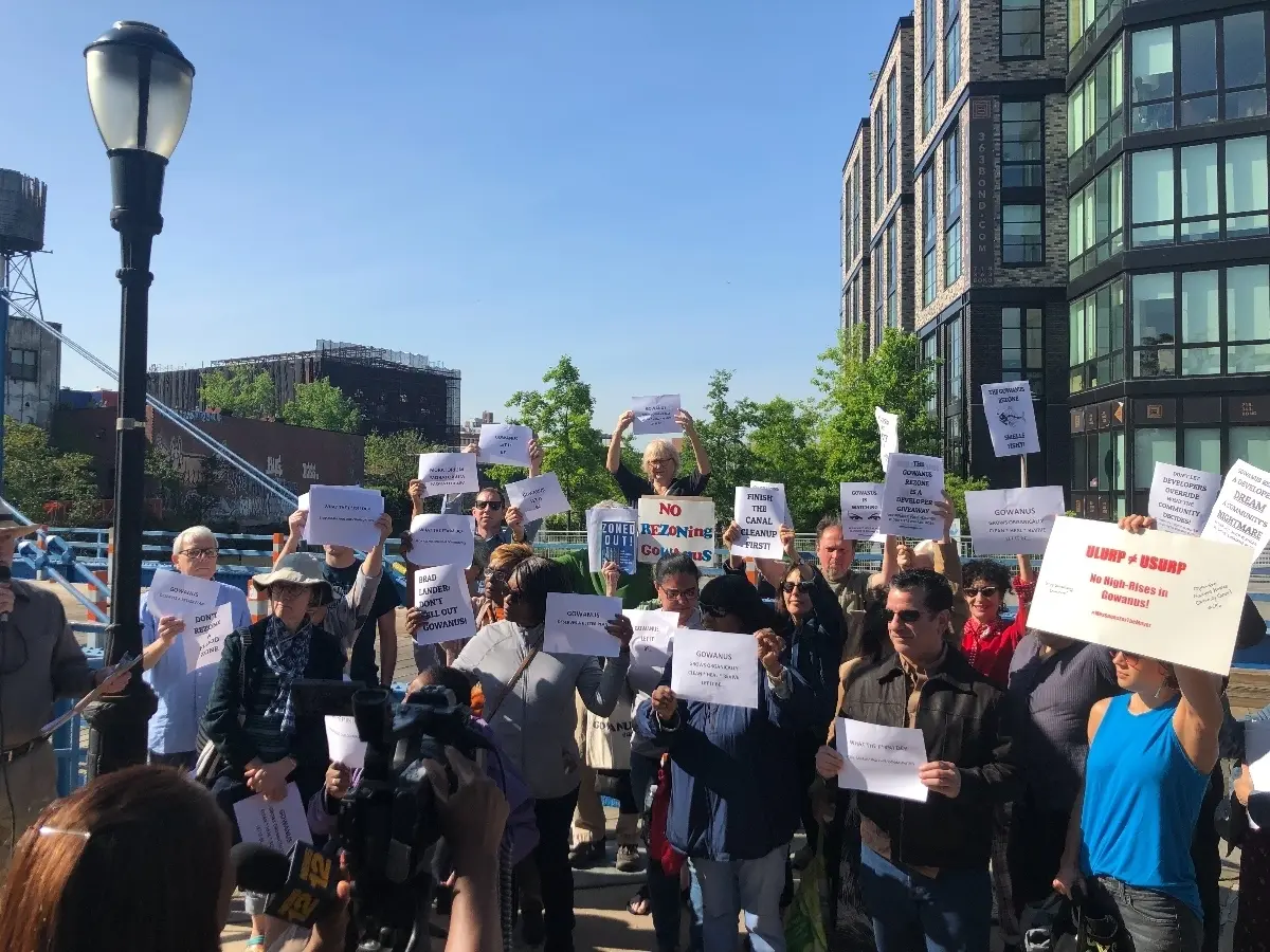 A crowd of Gowanus rezoning opponents holds protest signs outside a modern apartment building.